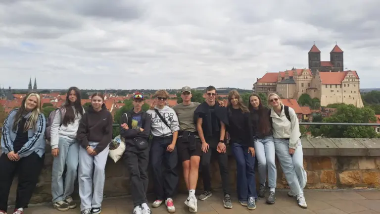 Grupa jauniešu stāv uz mūra sienas fona ar senu pili un pilsētu aizmugurē.