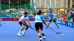 Sportisti sacenšas florbola spēlē uz zila laukuma āra apstākļos vasaras festivālā.