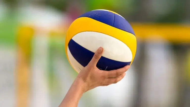 Rokas tur zilu, dzeltenu un baltu pludmales volejbola bumbu pret izplūdušu sporta laukuma fonu.
