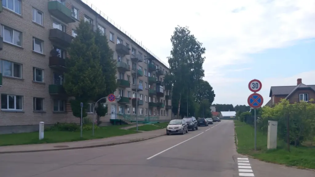 Daudzstāvu ķieģeļu dzīvojamā māja ielas malā ar automašīnām un ceļa zīmēm dienas laikā.