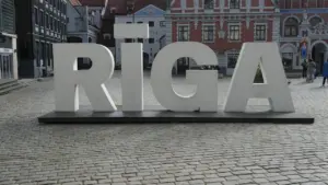 Liels, balts uzraksts RĪGA uz bruģēta laukuma Rīgas pilsētas centrā, fona ēkas.