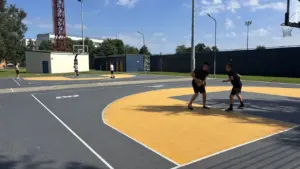 Vīrieši un pusaudži spēlē basketbolu uz āra laukuma ar pelēku un dzeltenu segumu.