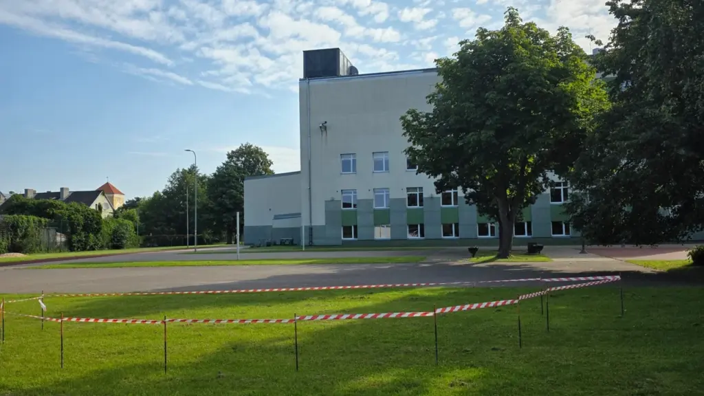 Zālienā apvilkta sarkanbalta brīdinājuma lente iepretim modernai administratīvajai ēkai saulainā dienā.