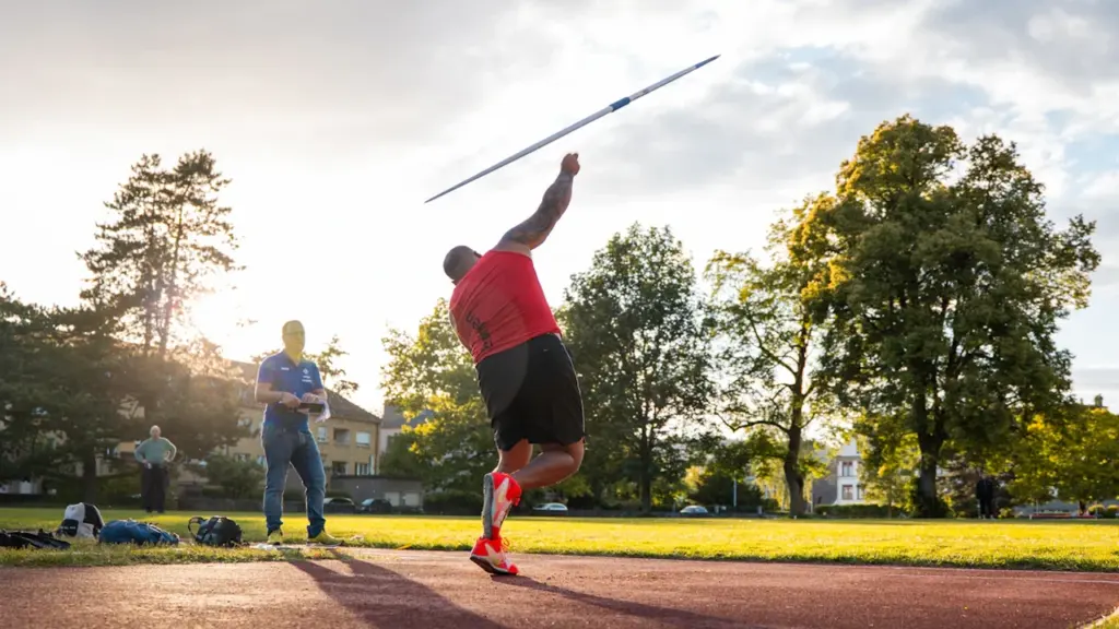 Sportists veic šķēpa metienu vieglatlētikas stadionā saulrieta gaismā, gatavojoties sacensībām.