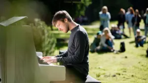 Jauns vīrietis tumšā džemperī spēlē baltas klavieres zaļā parkā ar klausītājiem fonā.