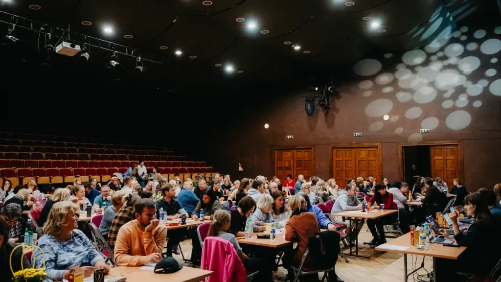 Cilvēku grupa sēž pie galdiem tumšā pasākumu zālē ar skatuvi un sarkanām krēslu rindām.